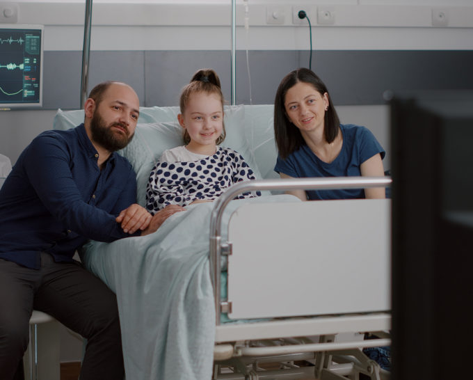 White little girl with her white parents sitting in a hospital bed watching TV.