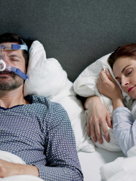 Man with sleep apnea wearing an oxygen mask in bed while a woman sleeps beside him.